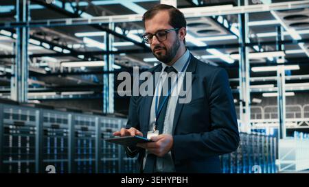 Data center computer scientist using tablet app, managing equipment powering online platforms. Man walks in server room, doing maintenance on digital storage systems using touchscreen device, camera B Stock Photo
