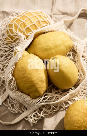 Overhead flat lay shot of fresh, vibrant yellow Italian lemons inside a reusable eco-friendly woven mesh string bag. The bag rests on a textured linen Stock Photo