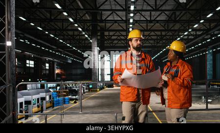 Diverse engineers reviewing manufacturing blueprints and technical drawings in heavy industry factory warehouse. Production hall with heavy machinery, instructions and layout plans. Stock Photo