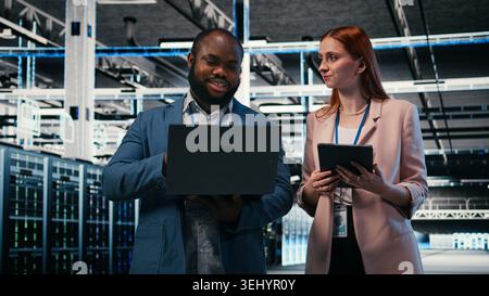 Server room teamworking colleagues using tablet and laptop to enhance operational effectiveness. Engineering team brainstorming in data center, using devices to monitor gear rackmounts, camera A Stock Photo