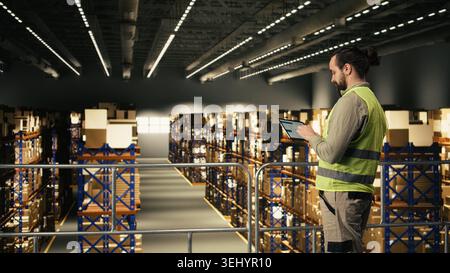 Male employee checking parcel status on a tracking software with tablet, shipment details and AWB tags ensuring package tracking in warehouse. Adult man wearing safety vest on platform. Stock Photo