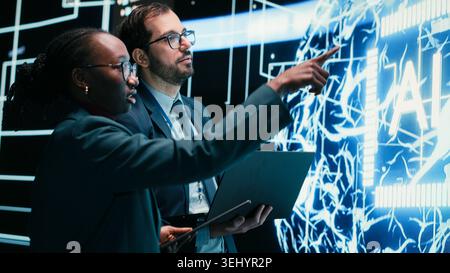 Engineering team coworkers receive tasks from autonomous agentic AI, working together. Teamworking colleagues talking with artificial intelligence superintelligence projected on digital wall, camera B Stock Photo