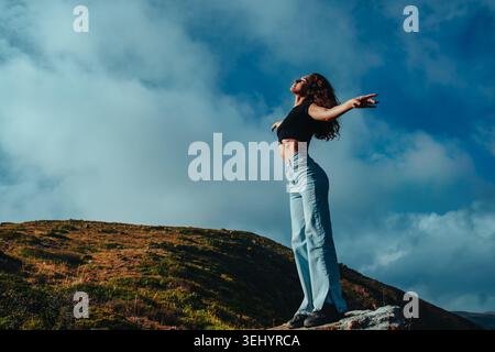 Brunette woman standing on rock with arms spread toward sun Stock Photo