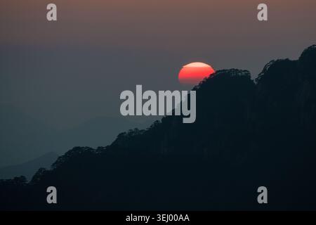 View of the sun setting behind the silhouetted peaks and pine trees of the Yellow Mountain in Huangshan City, Anhui, China. Stock Photo