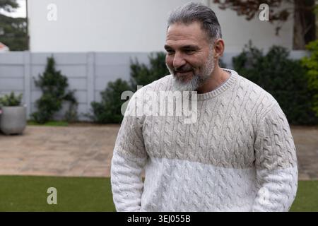Mature adult man standing on green turf in backyard wearing beige cable-knit sweater near planters Stock Photo