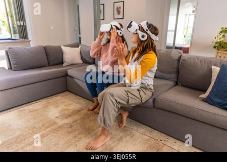 Two female friends using white VR headsets on gray sofa in living room, adjusting and clapping Stock Photo