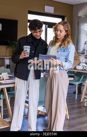 Diverse coworkers examining tablet in office, woman in denim jacket, man holding takeaway coffee Stock Photo