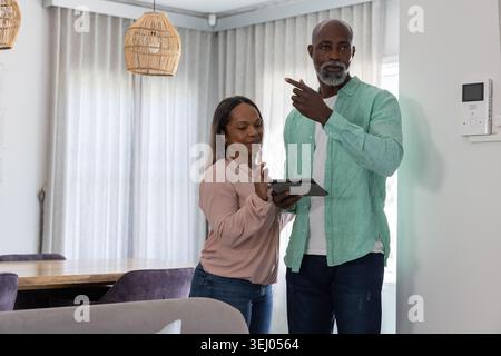 Mature adult couple standing near wall-mounted control panel in home, woman holding tablet Stock Photo