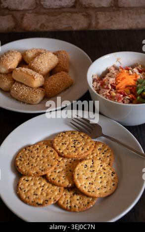 Plate with sesame seed crackers for snacking, on a white plate with tuna salad and vegetables, with a plate of buns filled with cream cheese in the ba Stock Photo