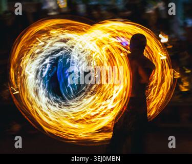 Person performing fire spinning with poi at night, creating abstract swirling light trails in vibrant orange, yellow, white, and blue hues, highlighting motion and energy Stock Photo