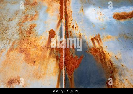 Heavily rusted metal surface with orange corrosion spreading across pale blue painted steel panel divided by central seam Stock Photo
