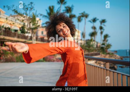 Beautiful woman joyfully raises her arms high celebrating life against a vibrant tropical background Stock Photo