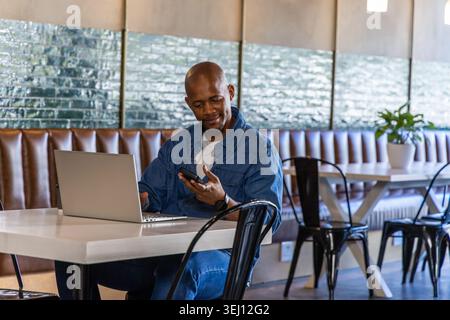 African man sitting at white table in cafe, wearing denim jacket, using laptop and phone Stock Photo