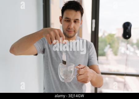 Portrait of smiling young man preparing cup of hot tea while window cleaning robot operates in background, savoring moment of relaxation Stock Photo