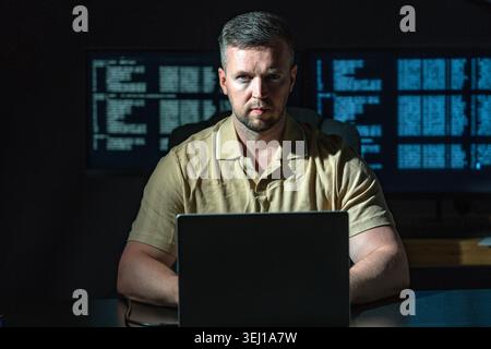 Hacker works in dark room alone. Hacker analyzes green code on laptop. Hacker monitors data flow in system. Hacker attempts network security breach. Stock Photo