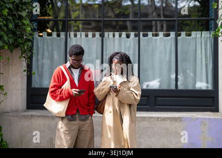 Thoughtful african american man and woman friends standing on street browsing online on smartphones. Stock Photo