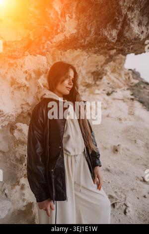 Woman leather jacket cliff standing against a sandy rock formation wearing a stylish white tracksuit during a warm sunset Stock Photo
