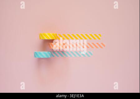 Three rolls of striped tape in yellow, orange, and blue unrolled on a pink background. Stock Photo