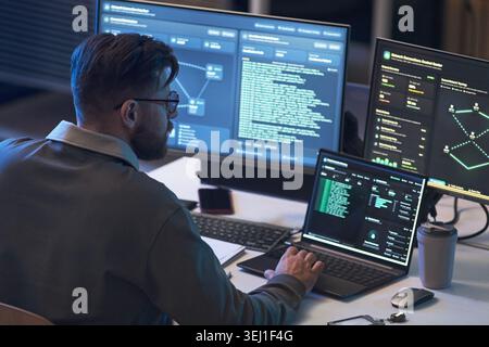 Young adult Caucasian man monitoring code and network dashboards across multiple screens, analyzing cybersecurity data. Ideal for IT security, software development, data protection marketing Stock Photo