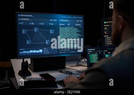 Caucasian young adult man monitoring network security dashboard and coding interface on multiple screens, analyzing cyber threat data. For cybersecurity, IT operations, data protection marketing Stock Photo