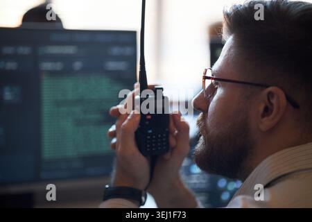Close up of Caucasian young adult man operating handheld radio beside computer monitors, coordinating communication in control room. Ideal for security, dispatch, operations content Stock Photo