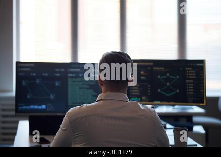 Rear view of young adult Caucasian man monitoring cybersecurity data on multiple screens in office, analyzing network activity and digital risk. Ideal for IT security, data protection marketing Stock Photo