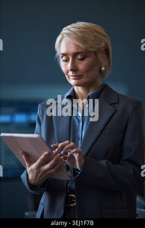 Vertical shot of middle aged Caucasian businesswoman using tablet, reviewing digital information in workplace. Suitable for corporate communication, leadership, digital workflow Stock Photo