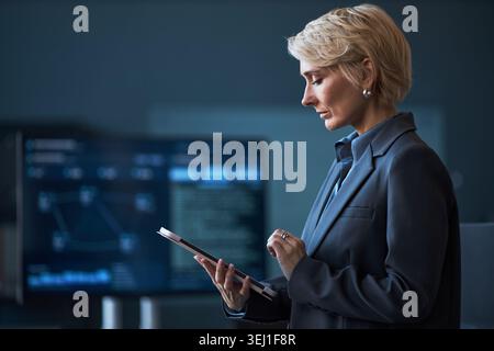 Medium shot of middle aged Caucasian businesswoman using tablet beside data screens, reviewing analytics in workplace. Ideal for digital strategy, leadership, corporate technology Stock Photo