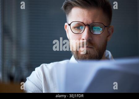 Close up of Caucasian young adult businessman reviewing documents through eyeglasses, focusing on office task and decision making. Suitable for business planning or finance content Stock Photo