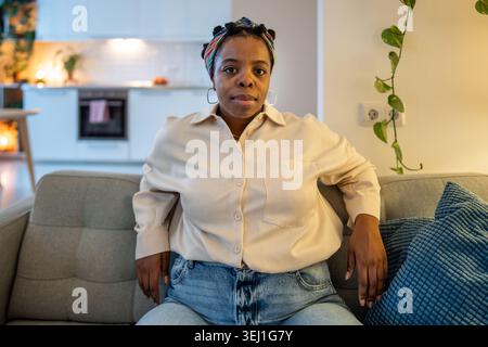 Self confident african american woman sits comfortably on couch looking at camera with calm expression. Self acceptance, body positivity, confidence, Stock Photo