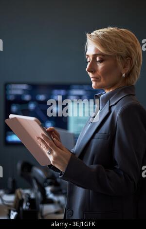 Vertical shot of middle aged Caucasian businesswoman using tablet while reviewing data in office, supporting digital workflow and executive decision making for business technology marketing Stock Photo