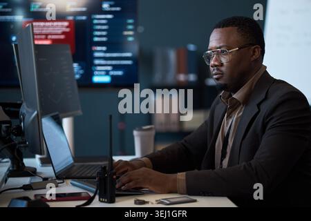 Black middle aged businessman monitoring cybersecurity data on desktop computer, working in control room. Ideal for IT security, network operations, risk management marketing Stock Photo
