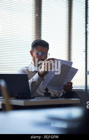 Vertical shot of Caucasian young adult businessman reviewing printed documents at office desk, checking paperwork and reports. Suitable for corporate finance, audit, compliance marketing Stock Photo