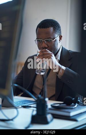 Vertical shot of African American young adult businessman drinking water while working at laptop, taking break during office task. Useful for workplace wellness, productivity, hydration Stock Photo