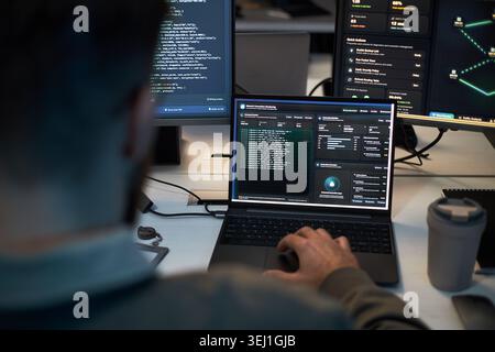 Caucasian man monitoring cybersecurity dashboard and code across laptop and multiple screens, analyzing network threats and system data. For IT security, data protection, SOC marketing Stock Photo