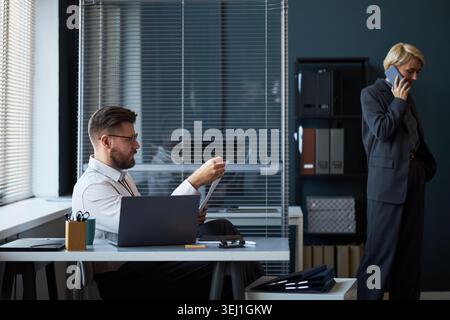 Caucasian businessman reviewing documents at desk while businesswoman speaking on smartphone in office, showing corporate workflow and teamwork. For business communication or office management Stock Photo