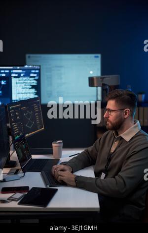 Vertical shot of Caucasian young adult man analyzing cybersecurity data on multiple screens and typing on laptop in office. Ideal for IT security, data analytics, tech workplace marketing Stock Photo