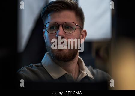 Caucasian young adult man wearing eyeglasses monitoring computer screens in office, conveying focus and data analysis. Useful for cybersecurity, IT support, digital work themes Stock Photo