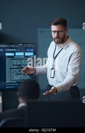 Vertical shot of Caucasian middle aged man presenting software code on monitor during team meeting, explaining cybersecurity workflow. Ideal for IT training, tech leadership marketing Stock Photo