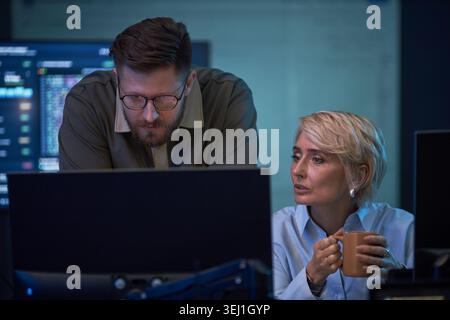 Caucasian coworkers reviewing data on desktop monitors in office, discussing problem solving and teamwork. Ideal for cybersecurity, IT support, software development, business operations Stock Photo
