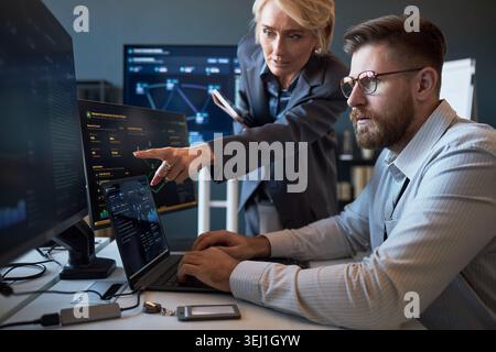 Caucasian businesswoman pointing at cybersecurity dashboard while Caucasian businessman analyzing data on laptop and monitors. Ideal for IT teamwork, network security, risk management Stock Photo
