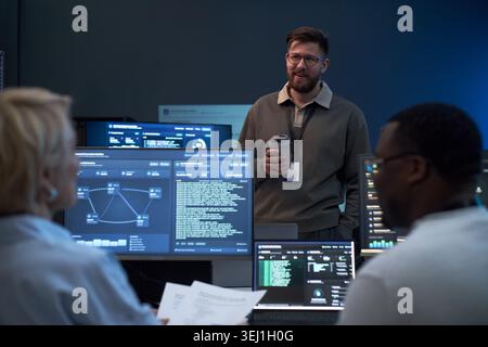 Medium shot of Caucasian young adult man leading cybersecurity briefing with diverse team, reviewing data on monitors. Ideal for IT operations, network security, workplace collaboration Stock Photo