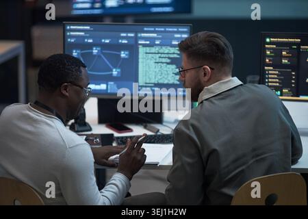 African American young adult man and Caucasian young adult man reviewing cybersecurity data on monitors, collaborating in office. For IT operations, network security, teamwork marketing Stock Photo