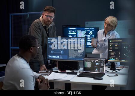 Multiethnic business team reviewing cybersecurity data on monitors, discussing network analysis in office. Ideal for IT operations, threat detection, digital security marketing Stock Photo