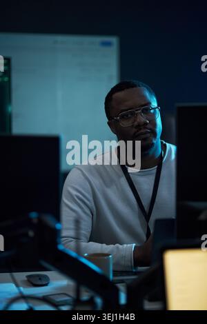 Portrait of Black middle aged man working at computer monitors in office, analyzing digital data and cybersecurity tasks. Ideal for IT services, tech staffing, network security marketing Stock Photo