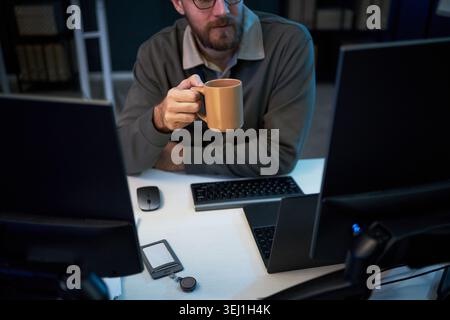 Middle aged Caucasian man holding coffee mug while working between multiple monitors and laptop at desk, suggesting late shift productivity. Ideal for remote work or tech workflow marketing Stock Photo