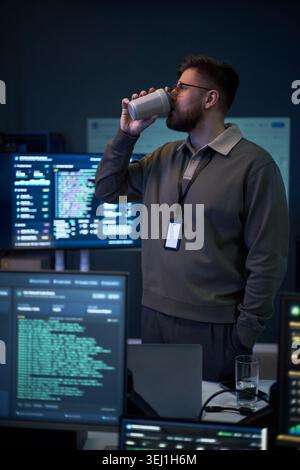 Vertical shot of Caucasian young adult man drinking coffee while monitoring code on multiple screens in tech office. Suitable for cybersecurity, software development, night shift work Stock Photo