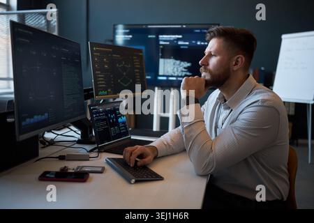 Caucasian young adult man analyzing cybersecurity data on multiple monitors in office, monitoring code and dashboards. Ideal for IT security, data analytics, tech workplace marketing Stock Photo