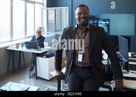 Portrait of African American young adult businessman standing in office, smiling at camera with colleague working behind. Ideal for leadership, corporate team, workplace branding Stock Photo