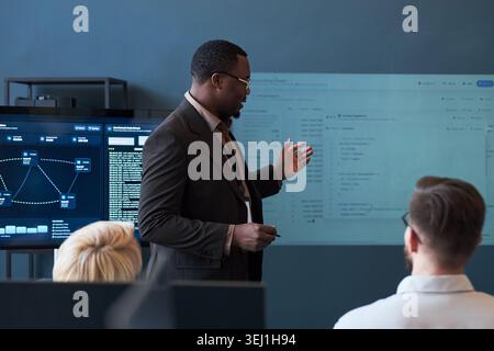 Medium shot of Black middle aged businessman presenting software dashboard to colleagues during office meeting, explaining data workflow. Ideal for business training, teamwork, IT strategy Stock Photo